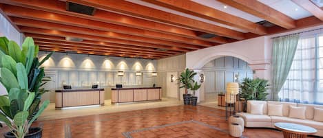 A spacious lobby with a wooden ceiling, a reception desk, a sofa, and potted plants.