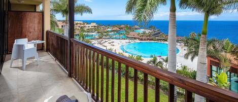 Balcón con vistas a la piscina y la playa de un complejo turístico, baranda de madera y una silla blanca.
