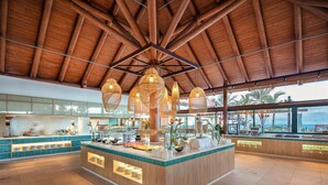 A spacious breakfast area with a wooden ceiling, a central island with food displays, and large windows offering a view of palm trees and the ocean. - Hotel La Palma Princess (Fuencaliente de la Palma)