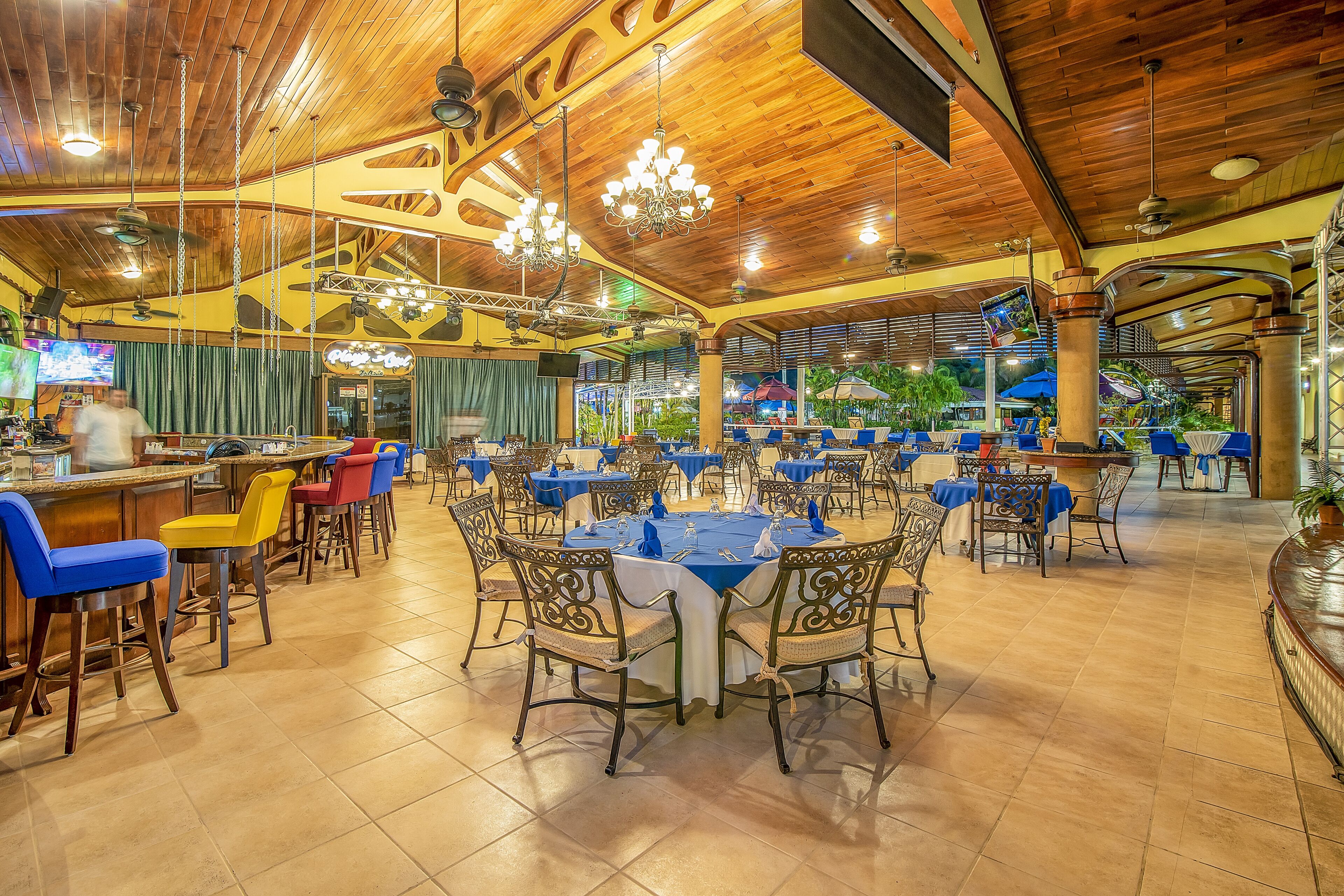 A spacious restaurant with high wooden ceilings, multiple dining tables with blue tablecloths, and a variety of chairs.