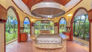 An outdoor kitchen with a brick island and a large overhead vent, surrounded by arches and a view of a tropical garden.