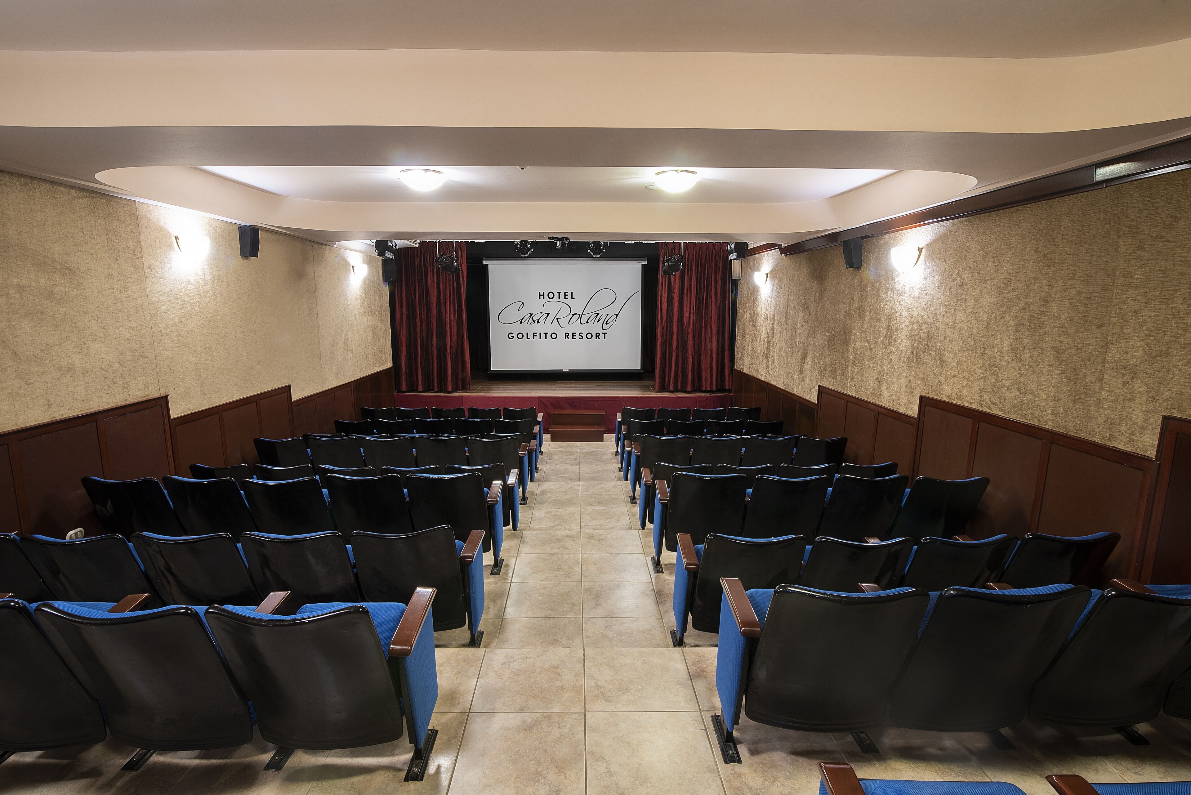 A theater with rows of black and blue chairs, a stage with a screen displaying 'Hotel CasaRocald Golfito Resort', and wooden paneling on the walls.