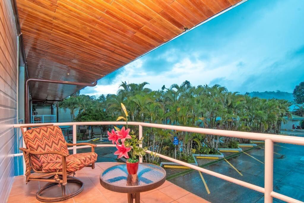 A balcony with a wooden ceiling, a rocking chair, a small table, and a potted plant with pink flowers.