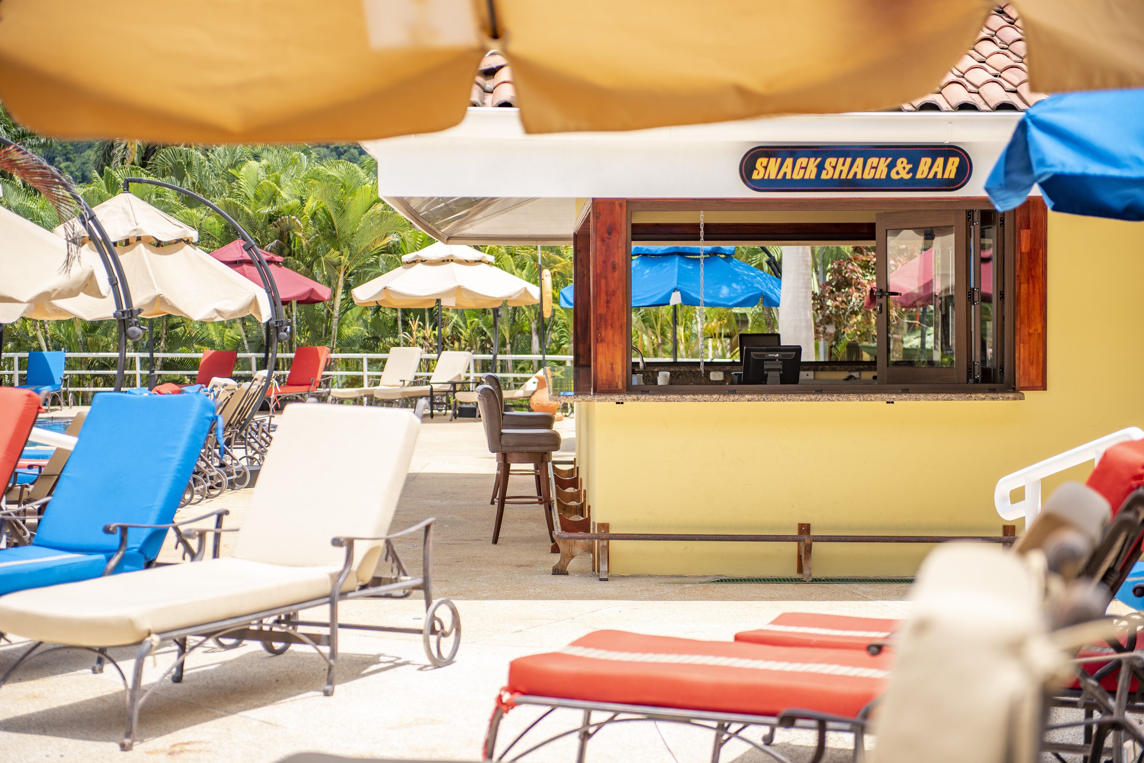 Un bar situé en bord de mer, avec une enseigne indiquant « SNACK SHACK & BAR », des tables et des chaises à l’extérieur, protégées par des parasols, et des fauteuils confortables.