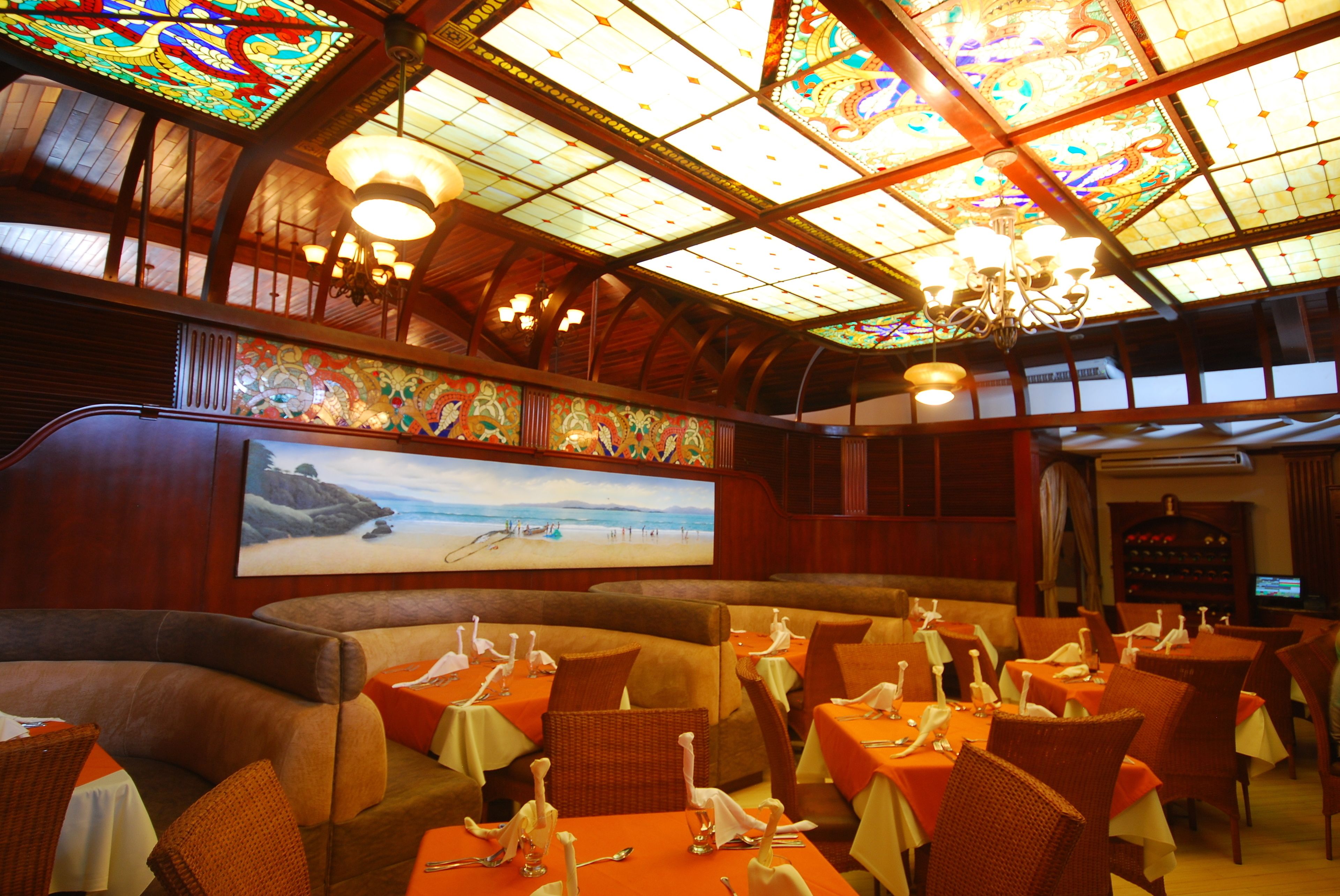 A restaurant with high ceilings, stained glass windows, and a large mural of a beach scene. Tables are set with white tablecloths and silverware.