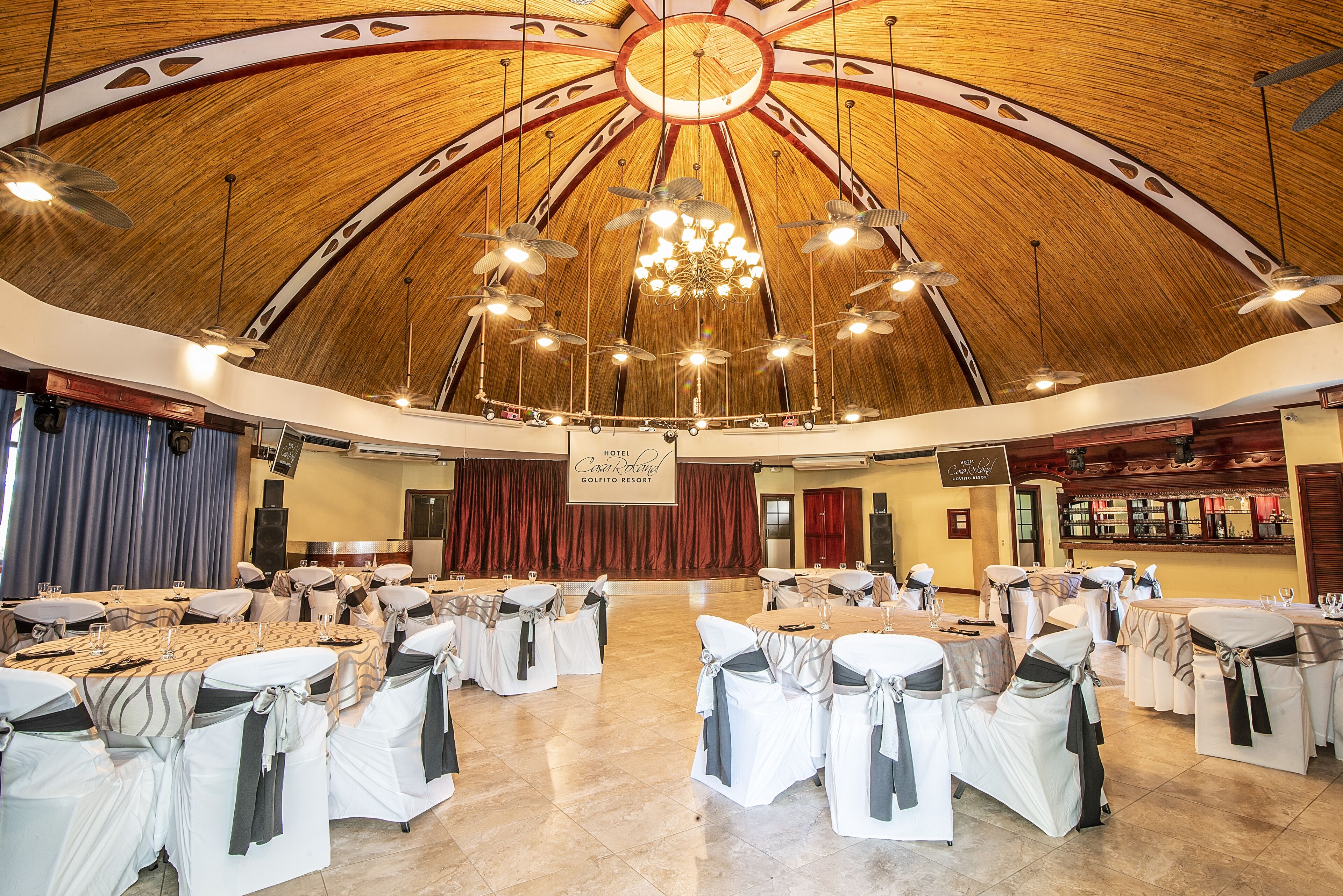 A large hall with round tables and chairs, a wooden ceiling, and a chandelier.