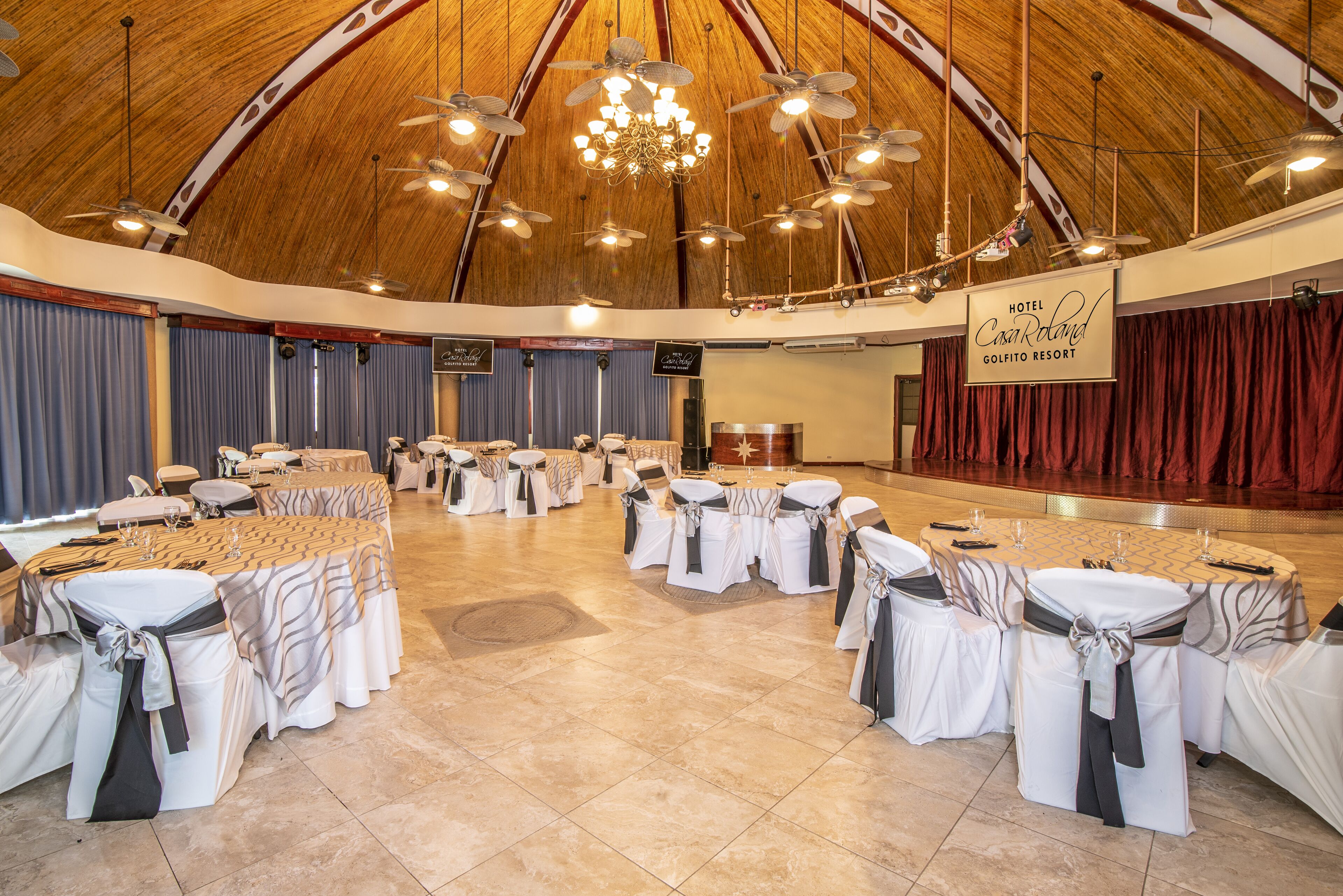 Une grande salle, semblable à une tente, avec de nombreuses tables et chaises rondes, une scène ornée d’une banderole et des ventilateurs de plafond.