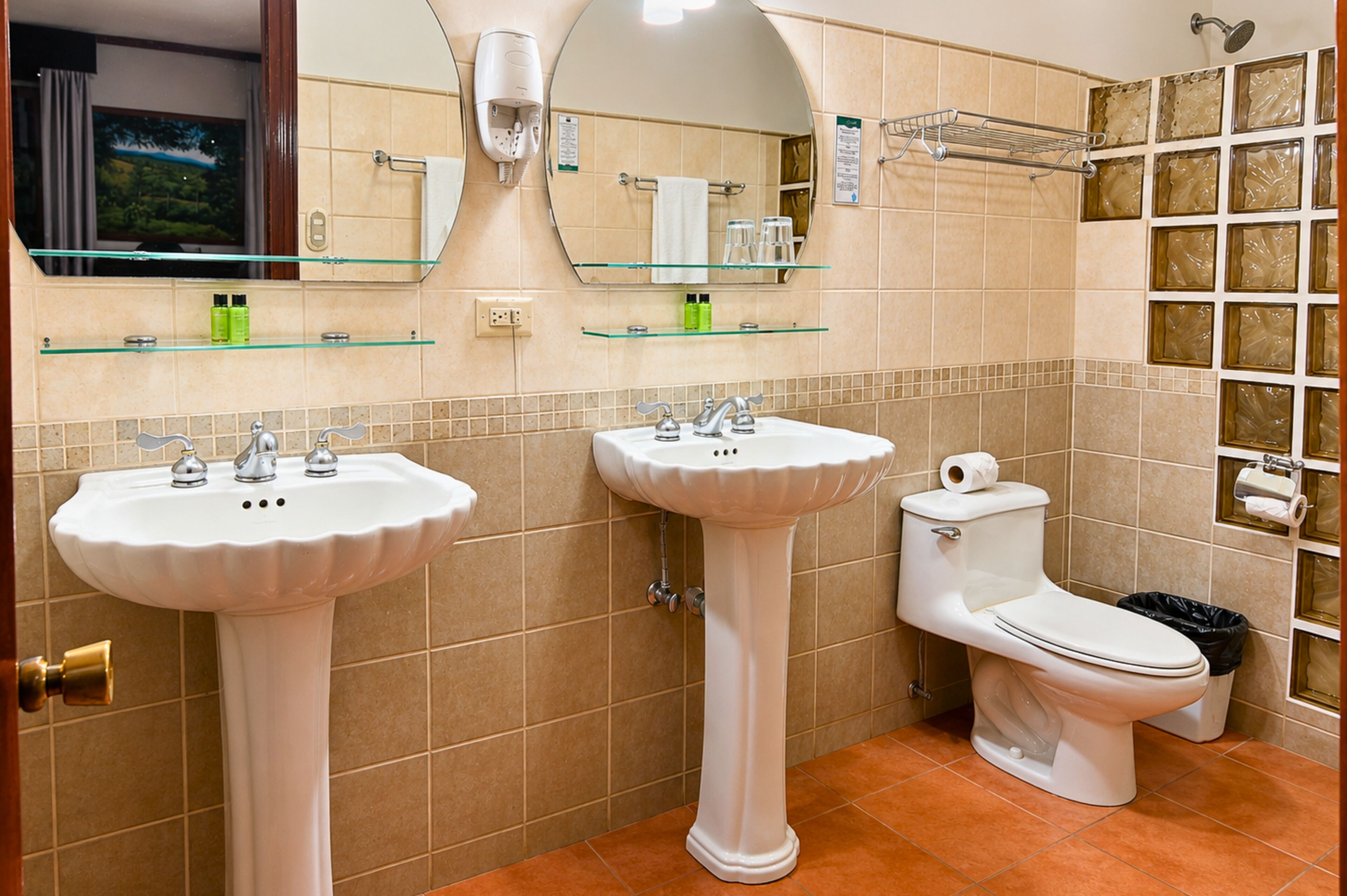 Un baño con dos lavamanos, un inodoro, un espejo y un estante de vidrio con artículos de aseo.