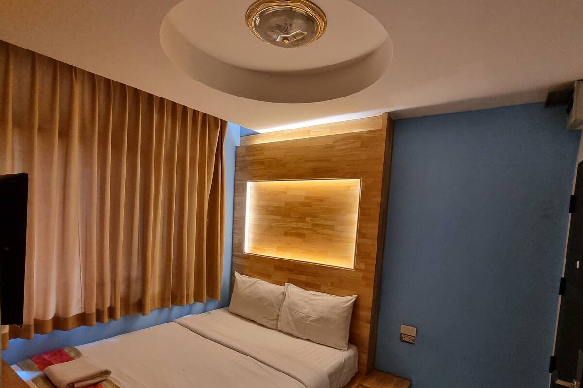 A modern hotel room with a wooden headboard, a large bed with white linens, a bedside table with a lamp, and a ceiling light fixture.