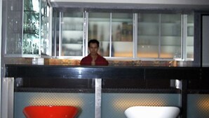 Zona de bar con una persona sentada tras la barra, dos taburetes con respaldo cónico y una ventana con reflejos.