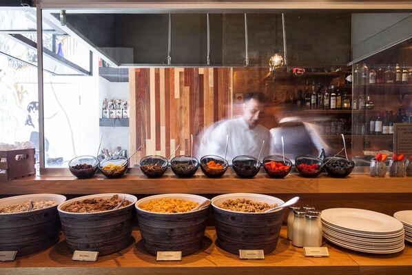Uma área de buffet com diversas opções de comida, incluindo tigelas de cereais e frutas, e um cozinheiro ao fundo.
