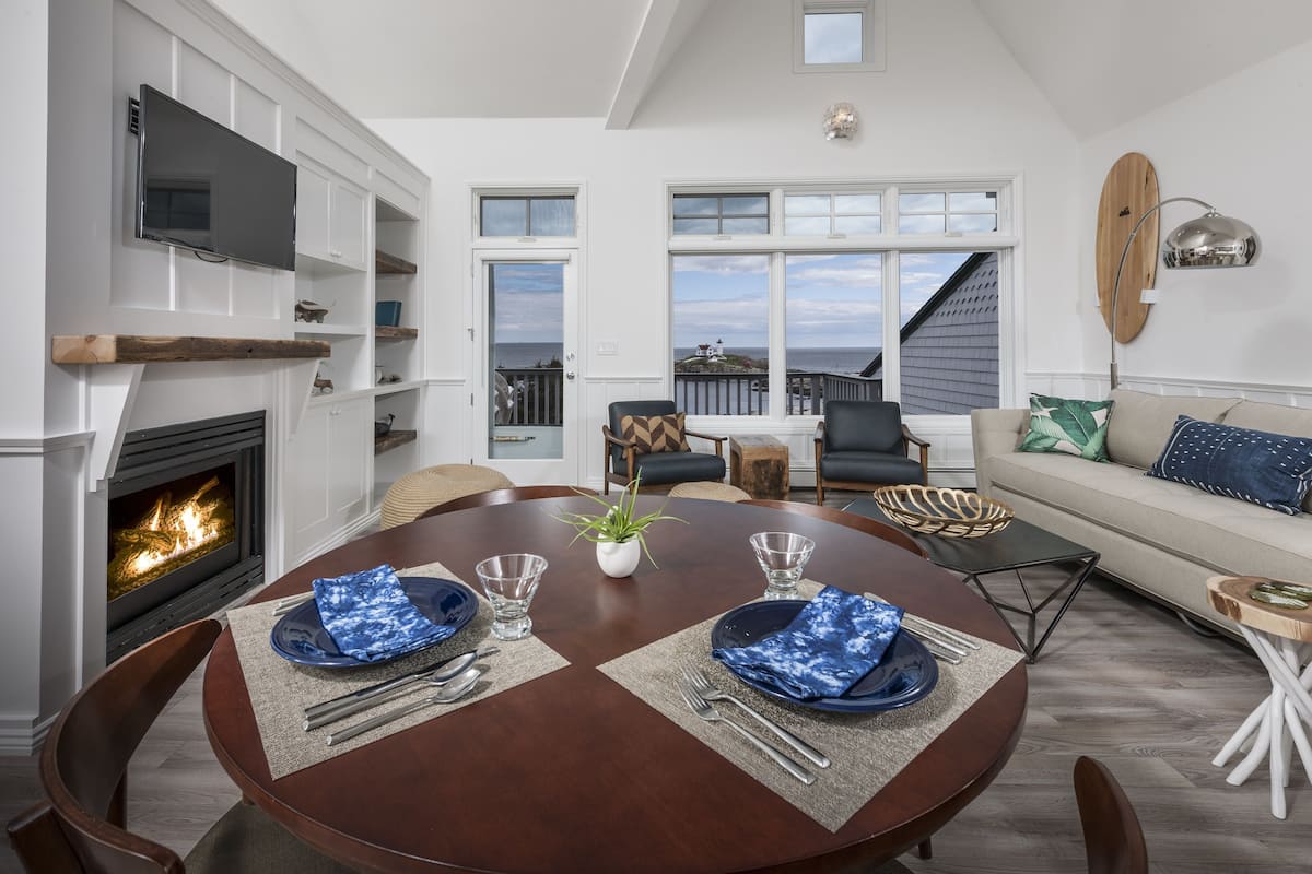 A dining area with a fireplace, a round table set for two, and a view of the ocean through large windows.