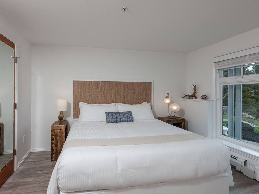 Uma cama grande com cabeceira de madeira, duas mesas de cabeceira com luminárias, uma janela com vista para o verde e uma porta com espelho.