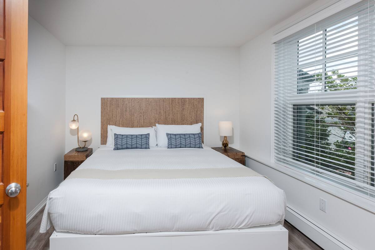 A neatly arranged bedroom with a large bed, two bedside tables with lamps, a wooden headboard, and a window with blinds.