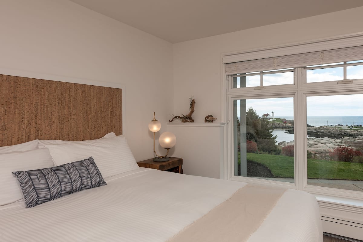 A bedroom with a large bed, a nightstand with a lamp, and a view of the sea and landscape through a window.