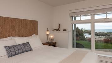 A bedroom with a large bed, a nightstand with a lamp, and a view of the sea and landscape through a window.