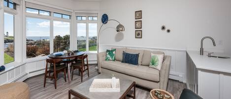 A modern living room with a dining area, large windows, a ceiling fan, and a kitchen area with a sink.