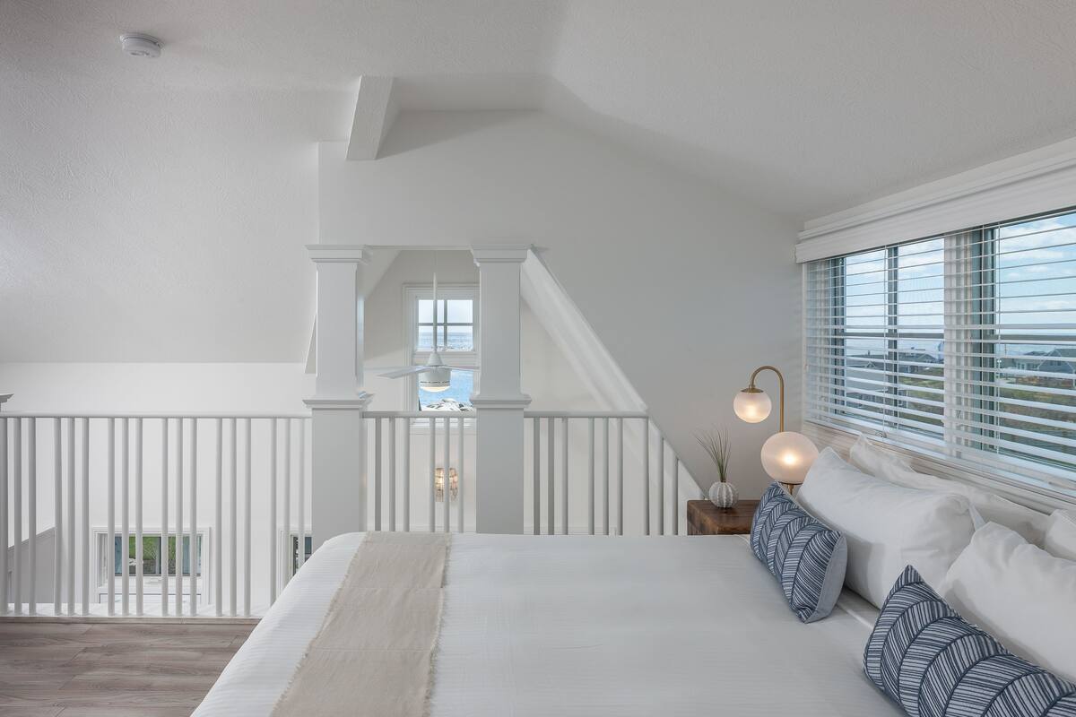 A bedroom with a large bed, white walls, a view of the outdoors, and a staircase leading to an upper level.