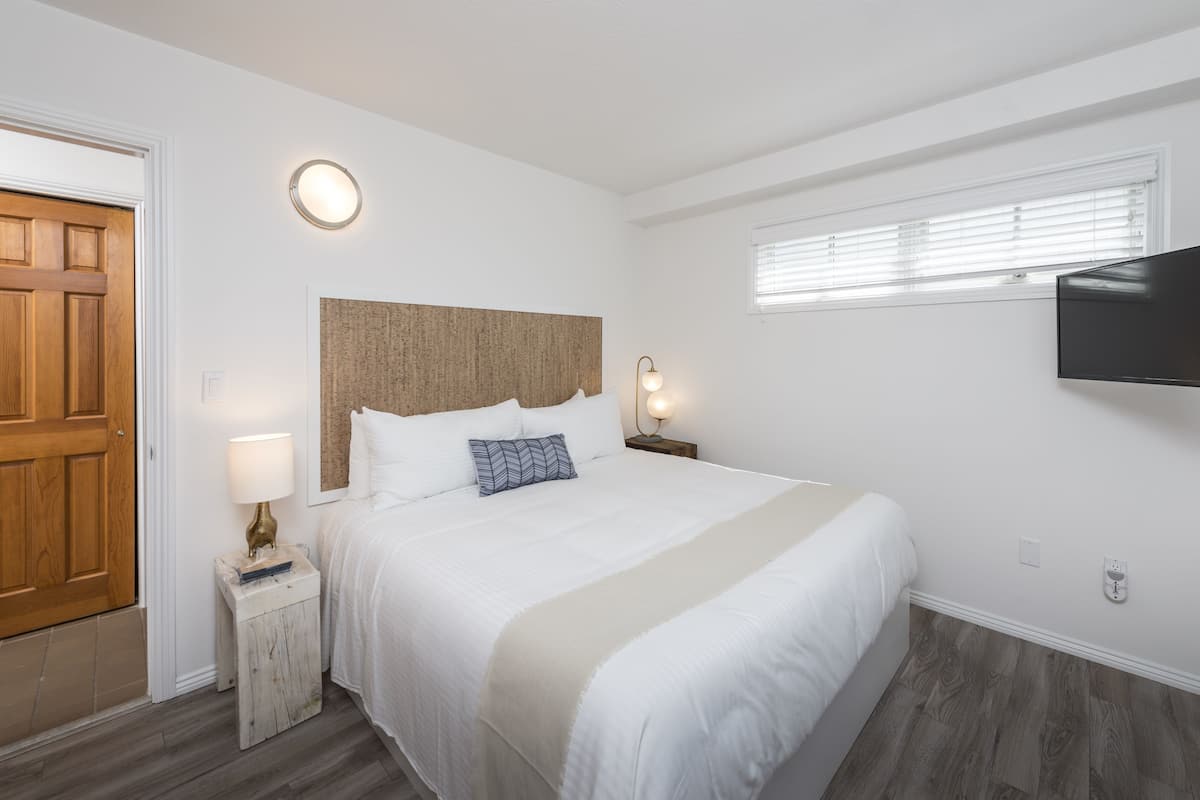 A bedroom with a wooden door, a bed with white bedding, a headboard, bedside lamps, and a mounted TV.