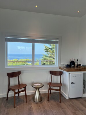 A room with two wooden chairs, a small round table, a white refrigerator, and a window with a view of the ocean and greenery.