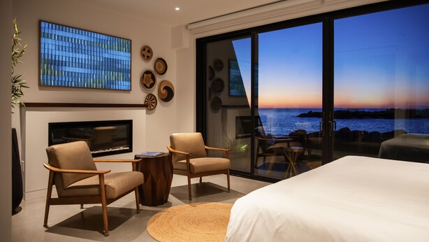 A modern hotel room with a large bed, two armchairs, a small table, and a fireplace. The room features a large window with a view of the ocean and a sunset. - ViewPoint Hotel (York Beach)