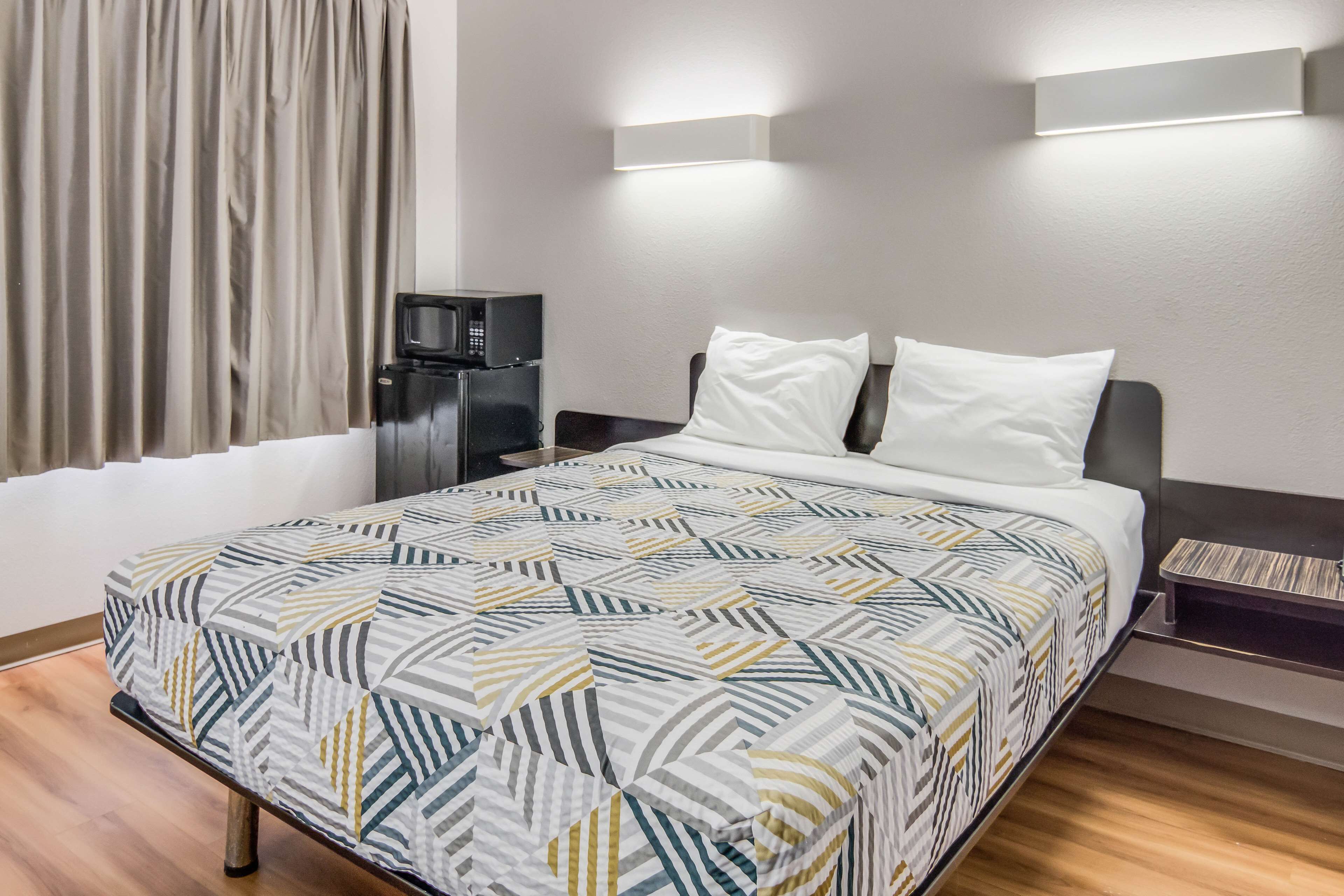 Una habitación de hotel con cama, mesita de noche, microondas e iluminación montada en la pared.