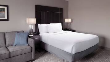 Una habitación de hotel moderna con una cama grande, un sofá gris, dos mesitas de noche con lámparas y un cuadro enmarcado en la pared.