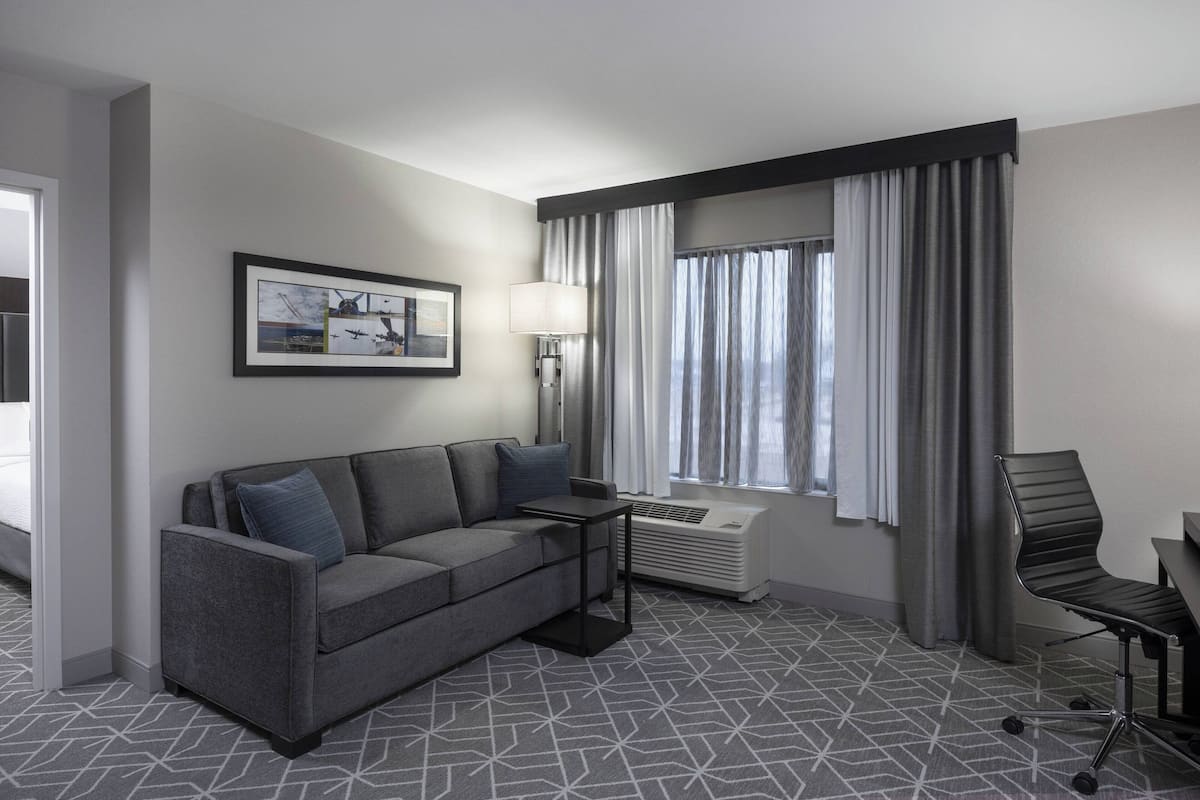 A modern hotel room with a grey patterned carpet, a grey sofa, a small table, a chair, a standing lamp, and a window with curtains.