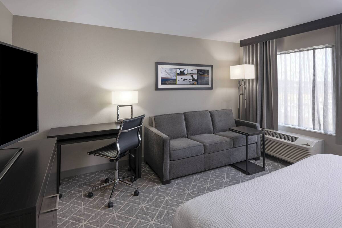 A hotel room with a bed, a desk with a chair, a grey sofa, a flat-screen TV, a framed picture on the wall, a lamp, and a window with curtains.