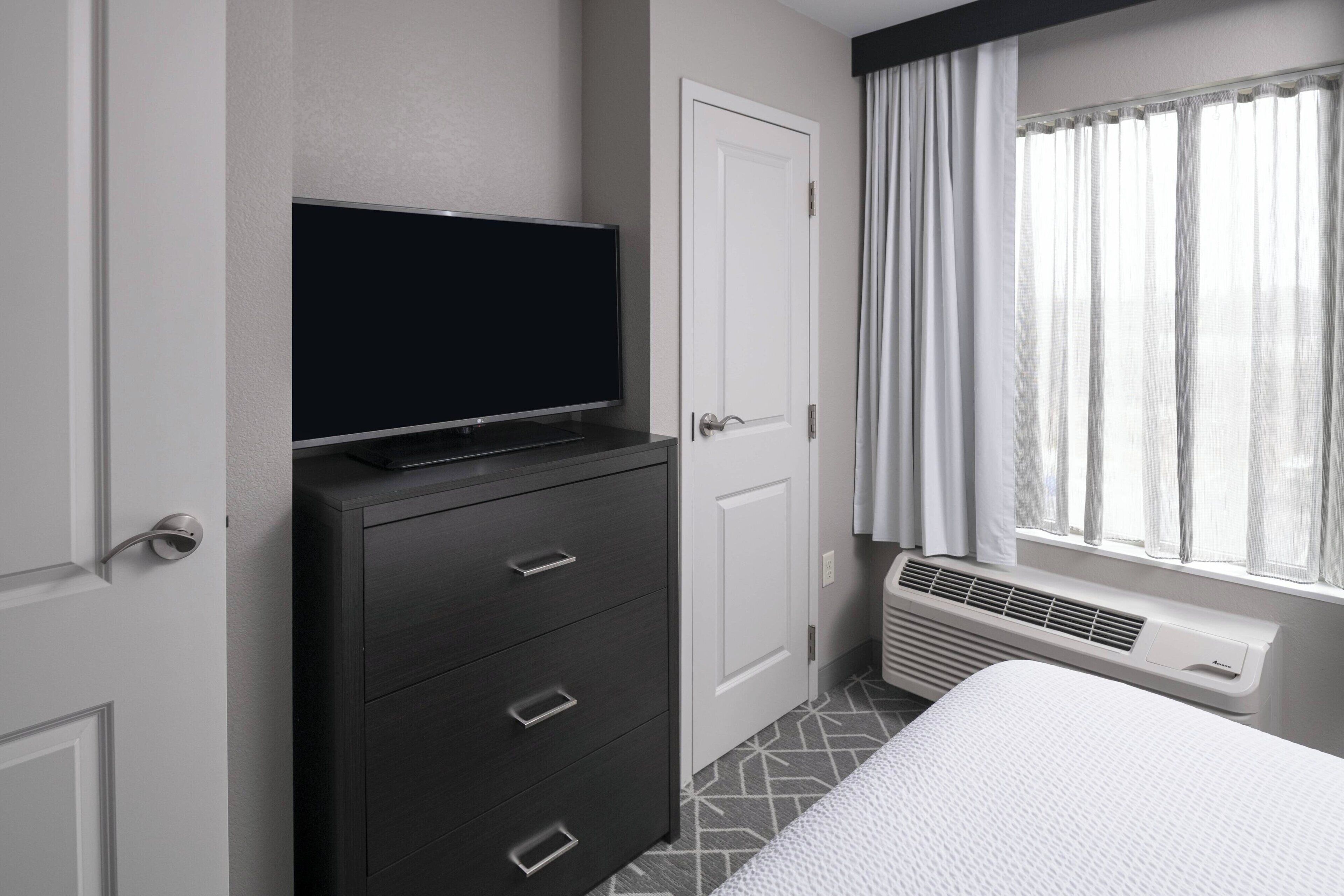 A hotel room with a flat-screen TV on a dresser, a bed with a white comforter, a window with sheer curtains, and a wall-mounted heater.
