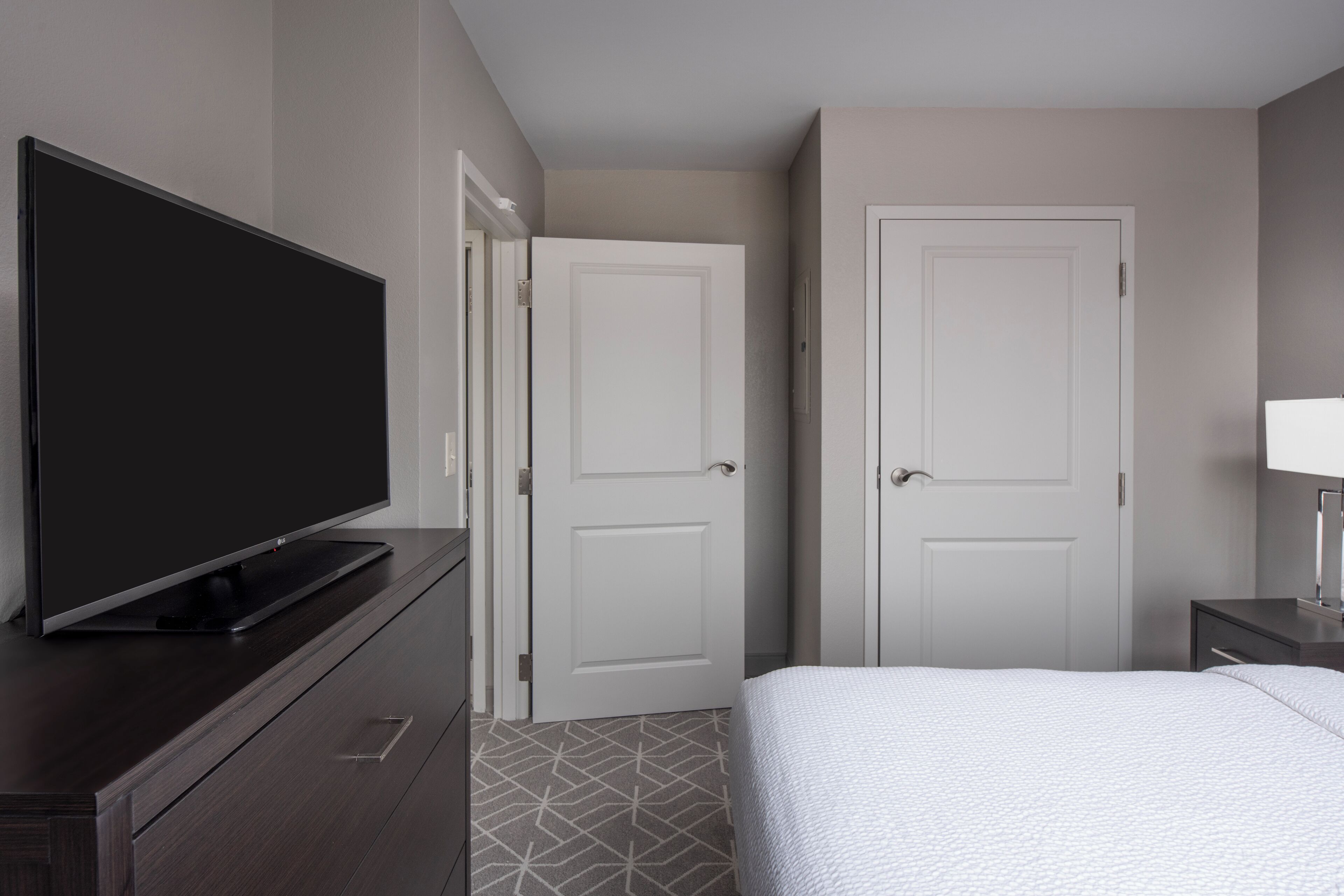 A hotel room with a flat-screen TV on a wooden dresser, two white doors, a bed with a white duvet, and a small bedside table with a lamp.