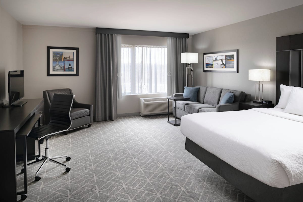 A hotel room with a large bed, a desk with a chair, a sofa, two wall-mounted lamps, and a window with curtains.