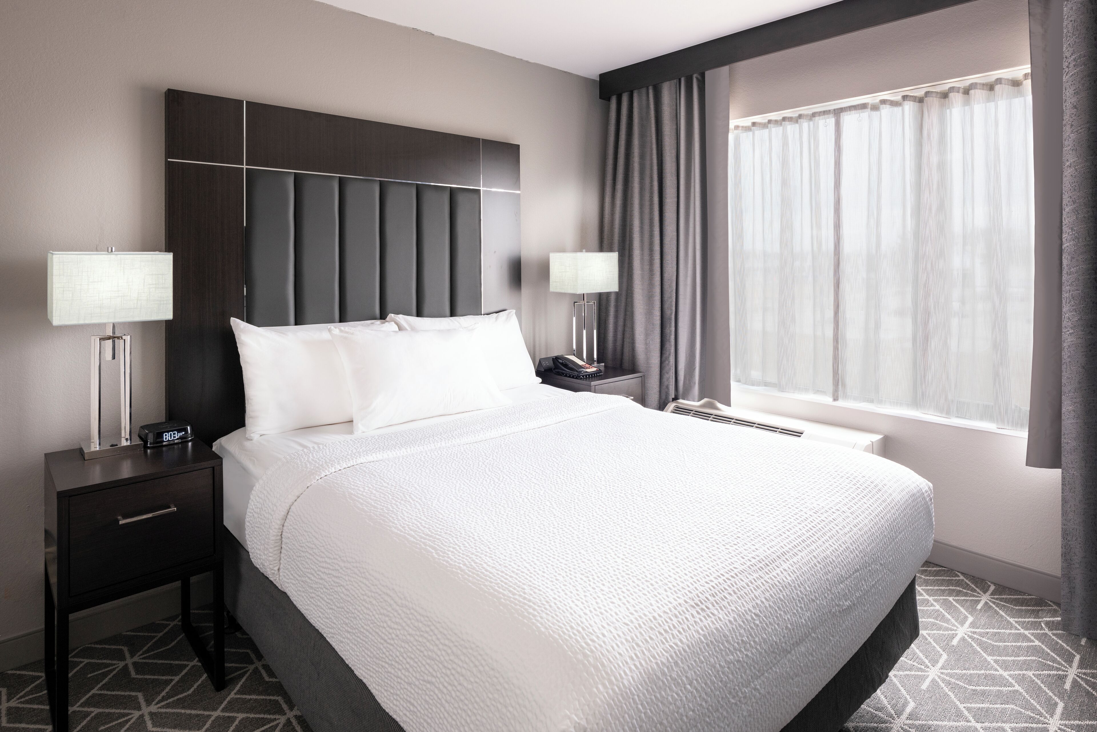A hotel room with a large bed, two bedside tables with lamps, a modern headboard, and a large window with curtains.