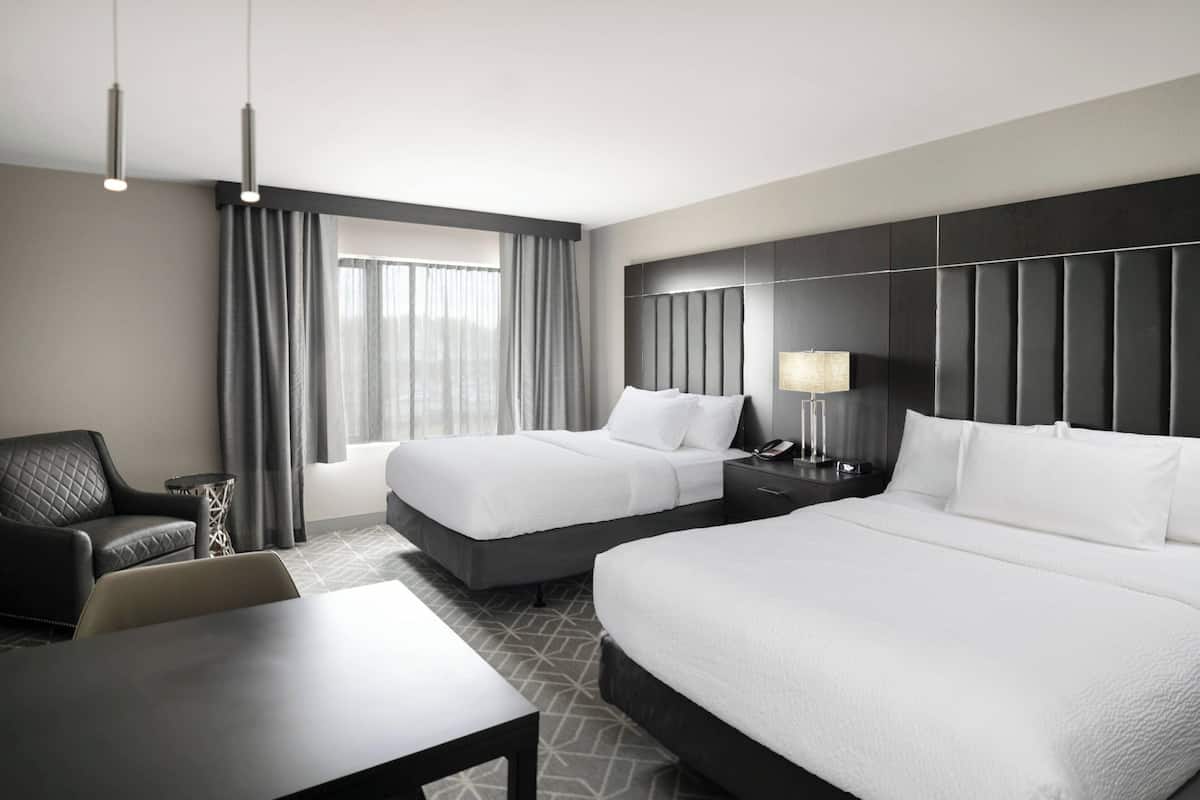 A hotel room with two beds, a desk, a chair, a lamp, and a view of the outdoors.