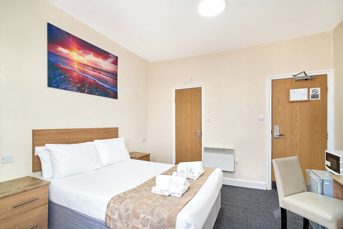 A hotel room with a bed, a wooden headboard, a painting of a sunset, a desk with a chair, and a door with a sign.