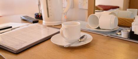 Escritorio de una habitación de hotel con una cafetera blanca, una taza de café, una cuchara y un menú en una tableta.