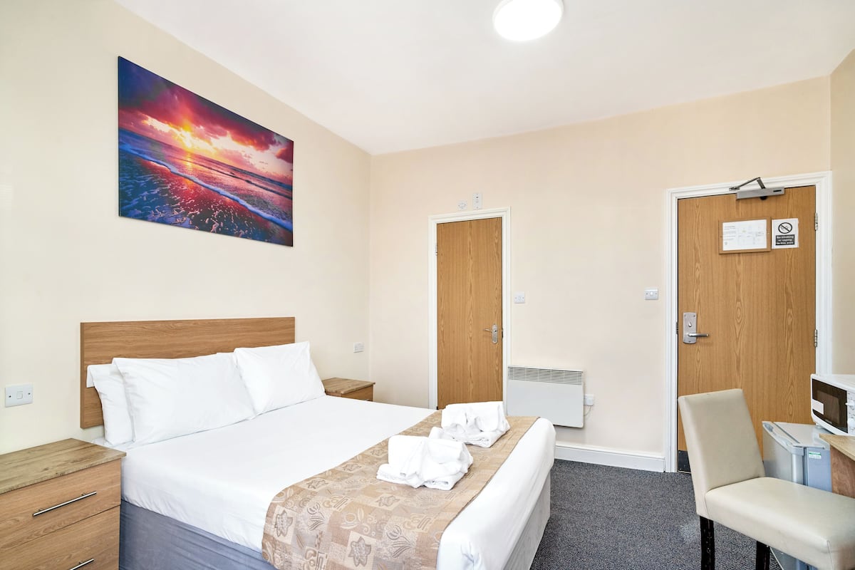 Una habitación de hotel con una cama, cabecera de madera, un cuadro de un atardecer, un escritorio con una silla y una puerta con un cartel.