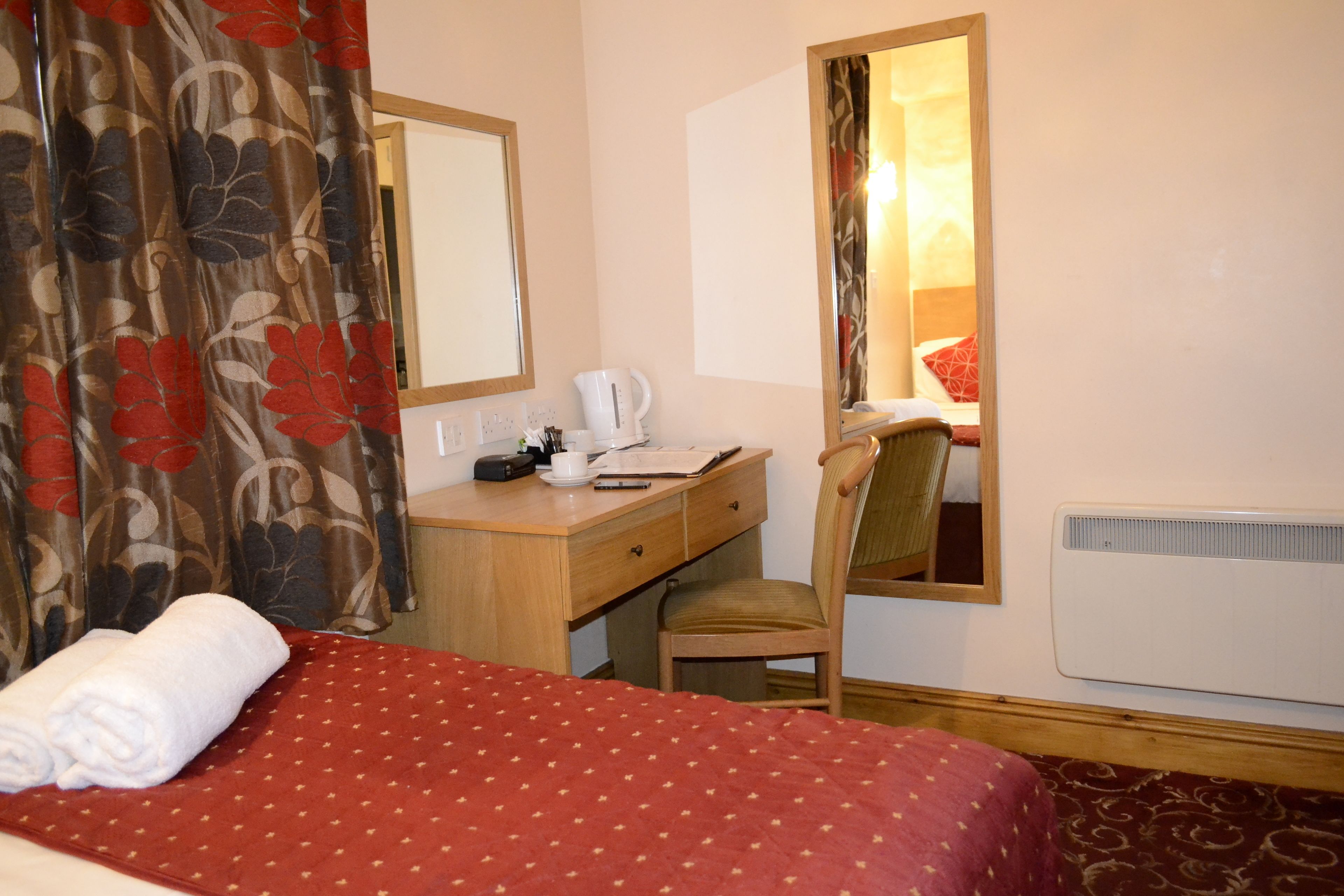 A hotel room with a bed, a desk, a chair, a mirror, a window, and a radiator.