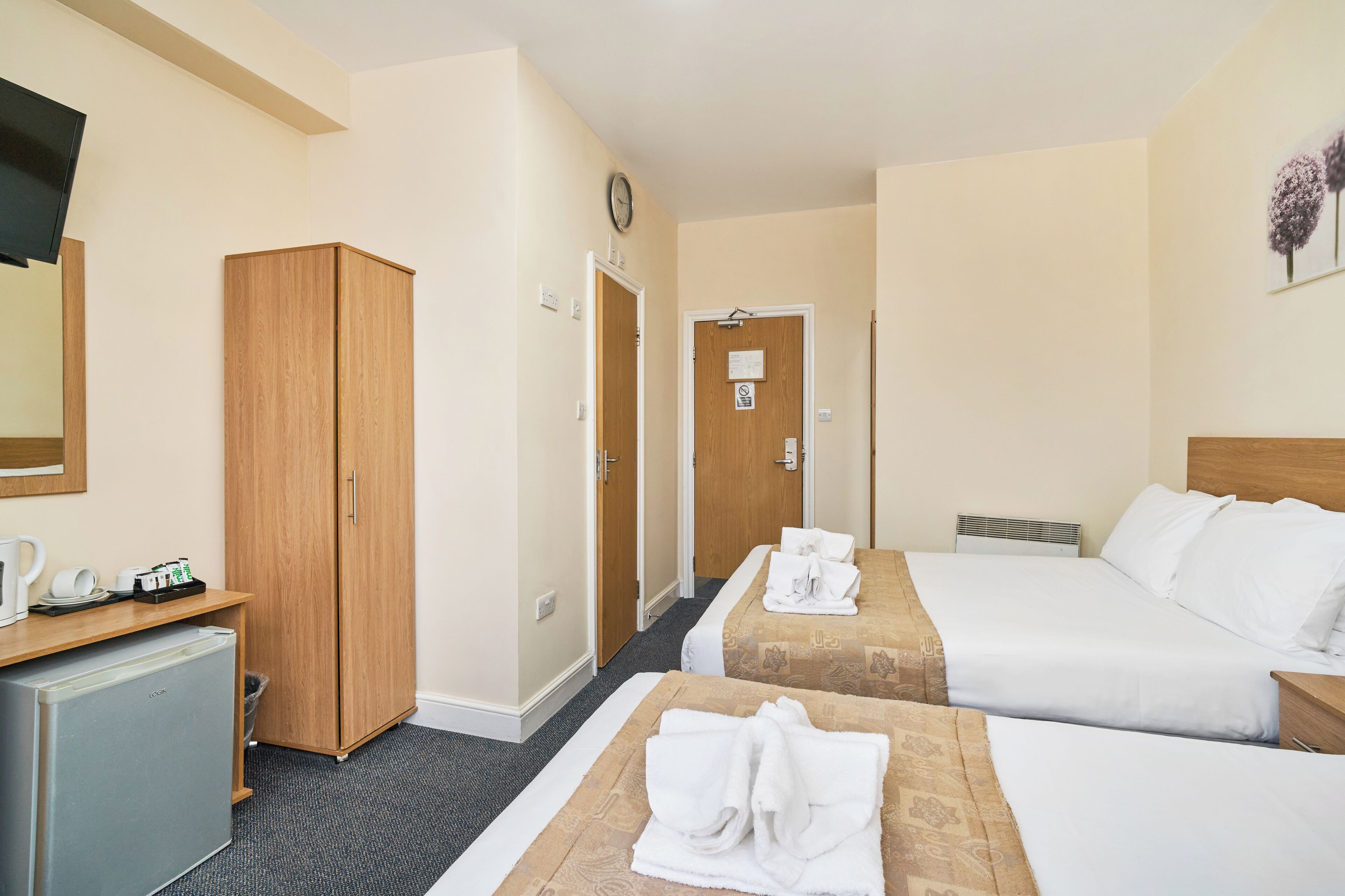 A hotel room with two beds, a desk with a refrigerator, a wardrobe, and a clock on the wall.
