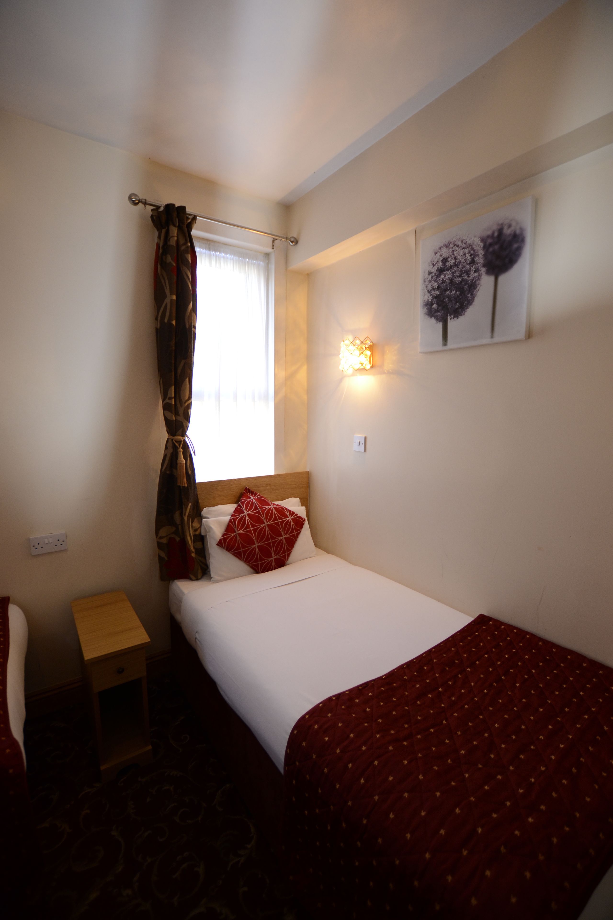 A hotel room with a single bed, a nightstand, a chair, a window with curtains, a wall-mounted light, and a painting.