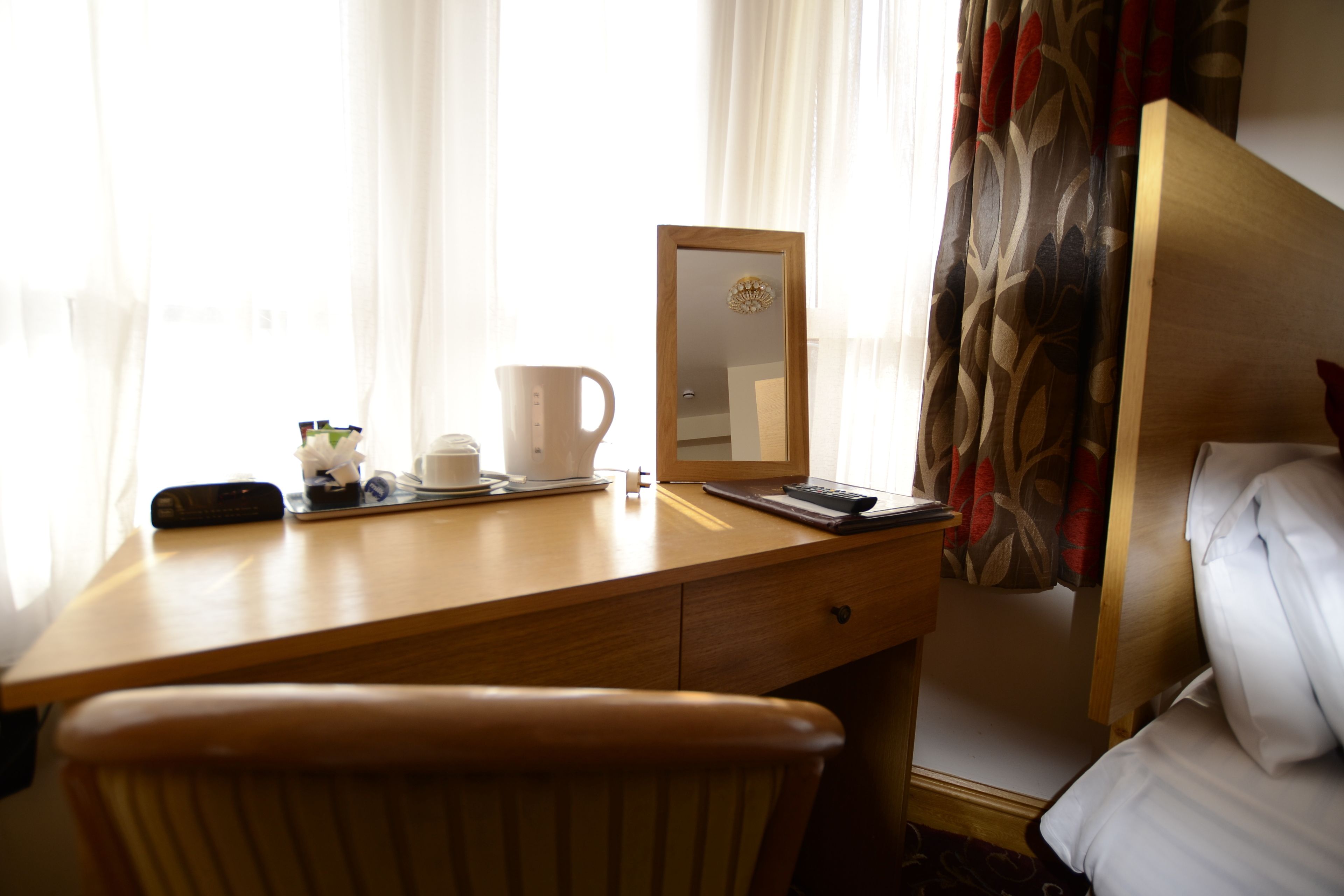 A hotel room with a wooden desk, a chair, a bed, a bedside table, a mirror, and a window with curtains.