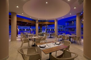 A spacious indoor dining area with multiple tables and chairs, a high ceiling with circular lighting, and a view of a building with balconies. - La Marquise Luxury Resort Complex (Rhodes)