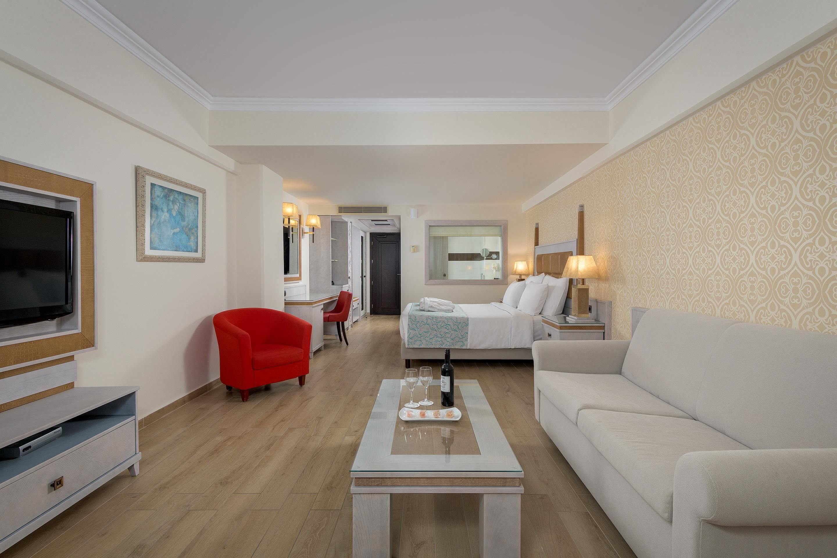 Une chambre d’hôtel avec une télévision, un fauteuil rouge, un canapé blanc, une table basse avec une bouteille et des verres, et un mur à motifs.