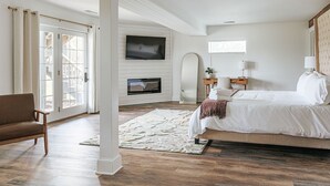 Un dormitorio con una cama grande, un televisor montado en una pared blanca, piso de madera y un ventanal amplio.
