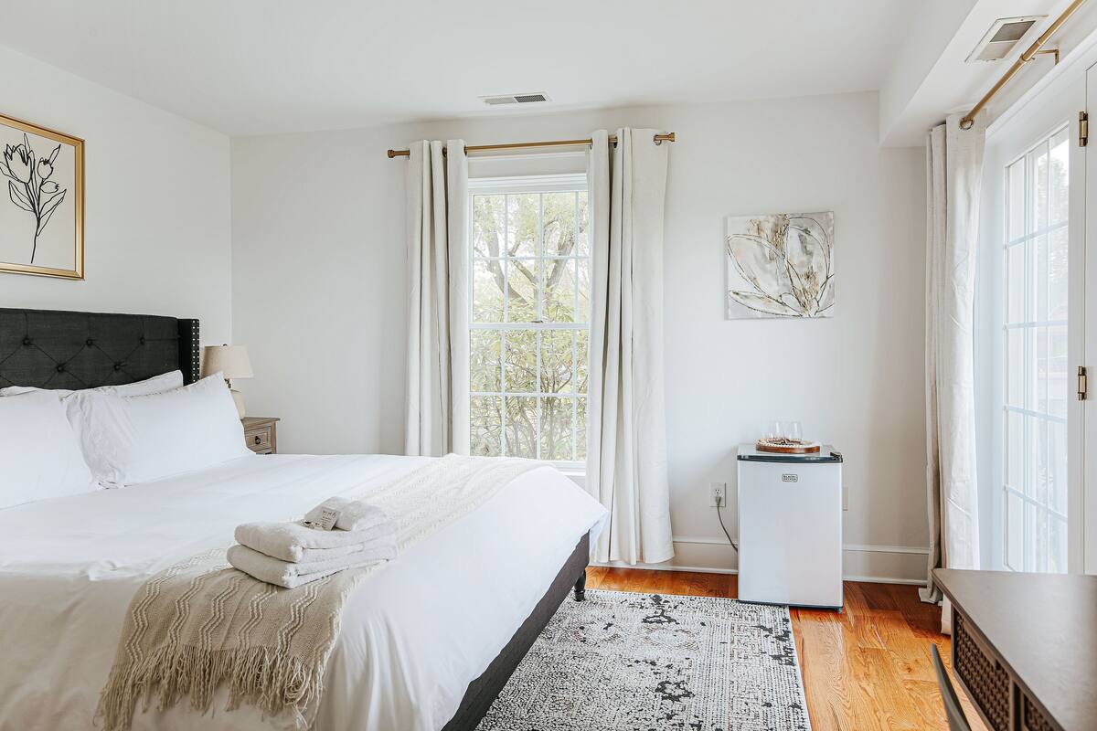 Quarto com cama, mesa de cabeceira, abajur, janela com cortinas, geladeira e tapete.