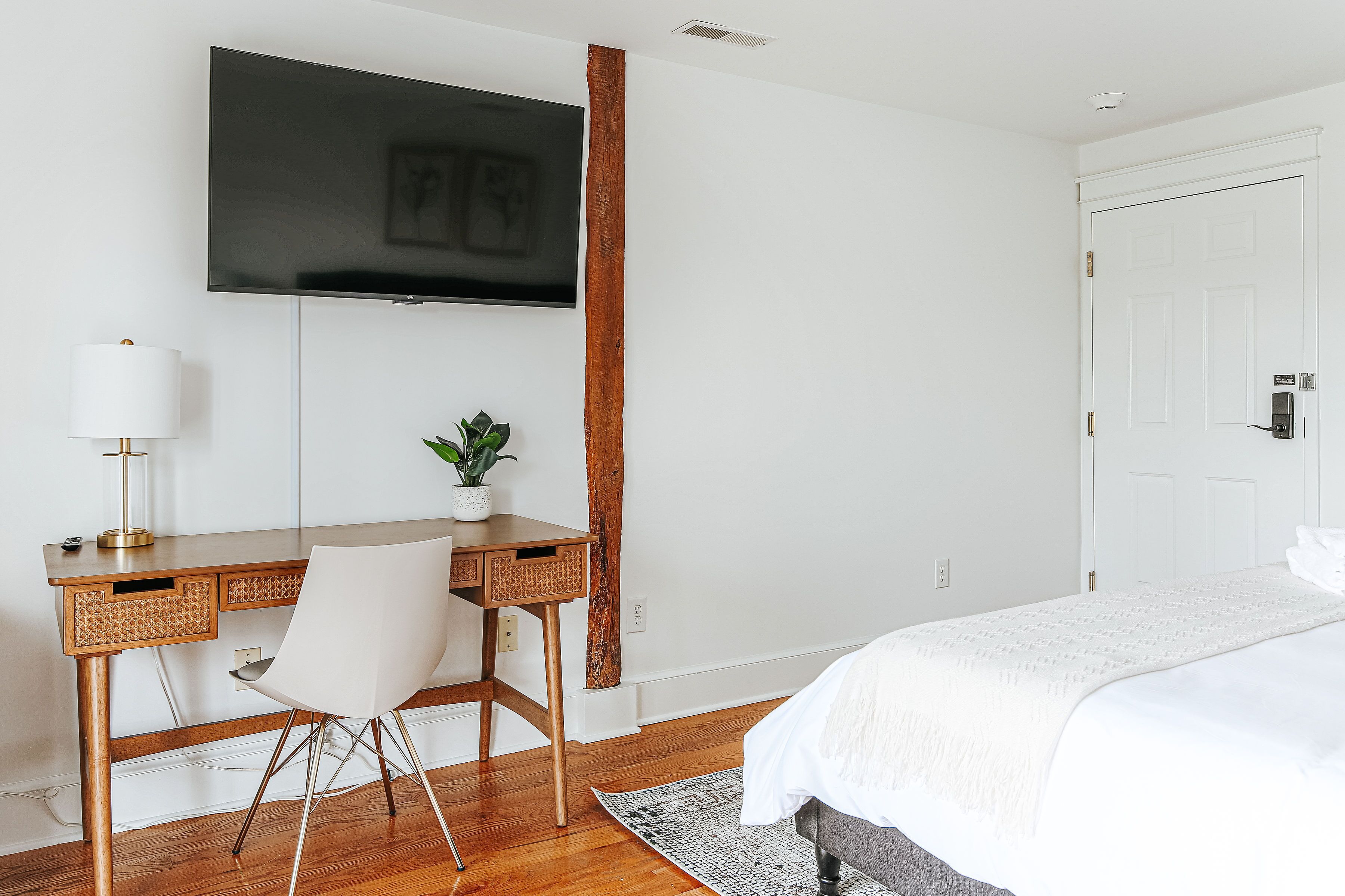 Quarto de hotel com uma escrivaninha de madeira, uma cadeira branca, uma cama com roupa de cama branca, uma televisão de tela plana fixada na parede e um pequeno vaso com planta na escrivaninha.