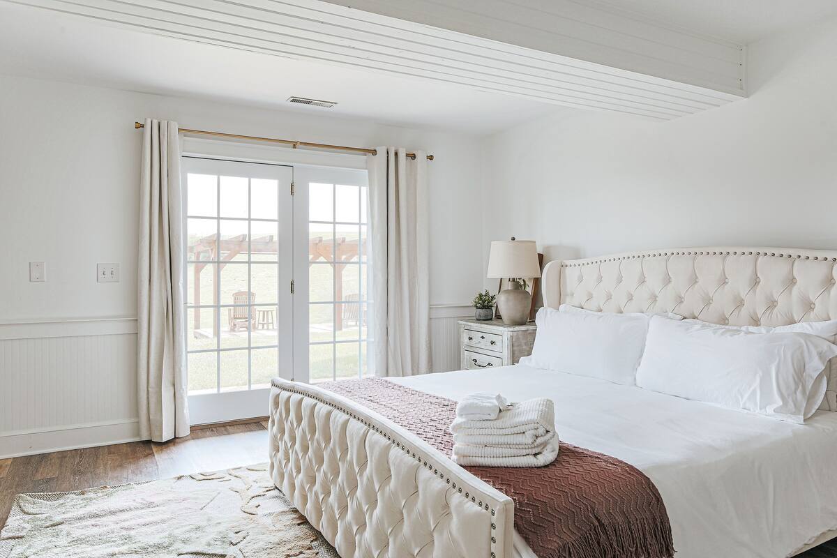 Quarto com cama grande, mesa de cabeceira com luminária e vista através de uma porta de vidro.