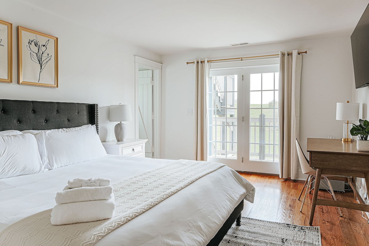 Cama bem arrumada com lençóis brancos e uma toalha dobrada, uma escrivaninha de madeira com abajur e vista para uma varanda através de uma porta de vidro.