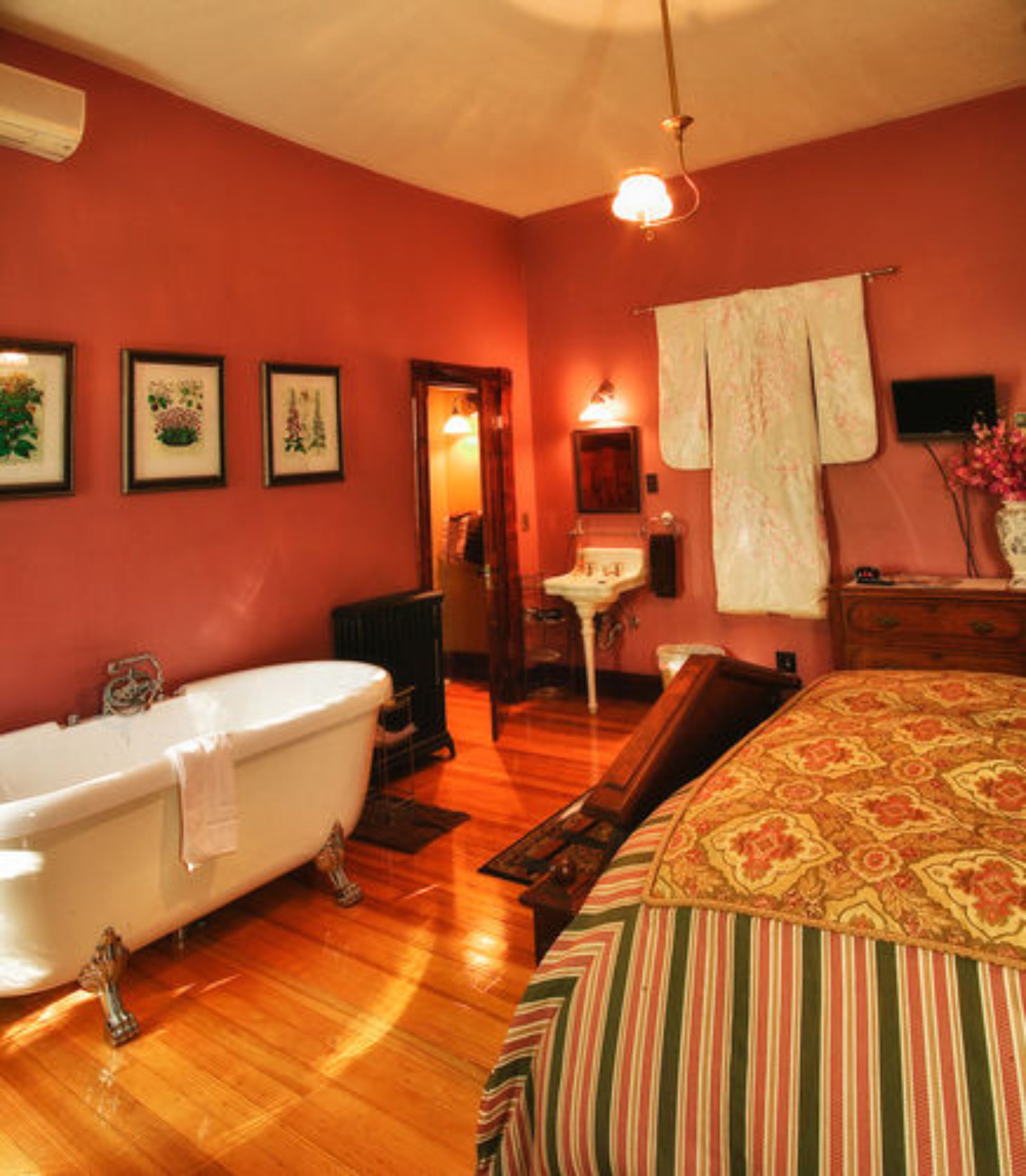 Une chambre avec une baignoire, un lit recouvert d’une courtepointe à rayures, une commode en bois, une coiffeuse avec un miroir et des tableaux encadrés accrochés au mur.