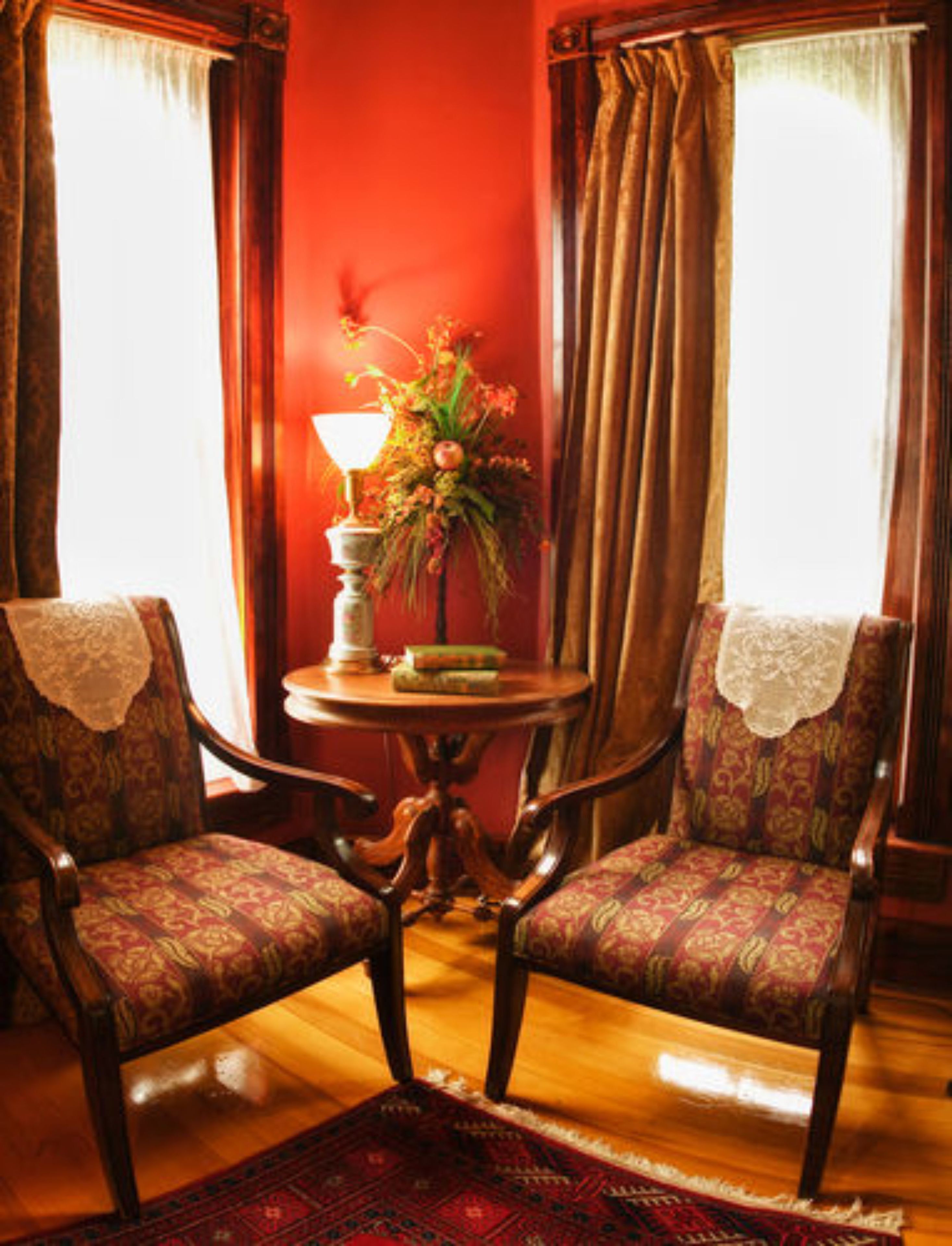 A room with two upholstered chairs, a round wooden table with a lamp and a floral arrangement, and a red wall.